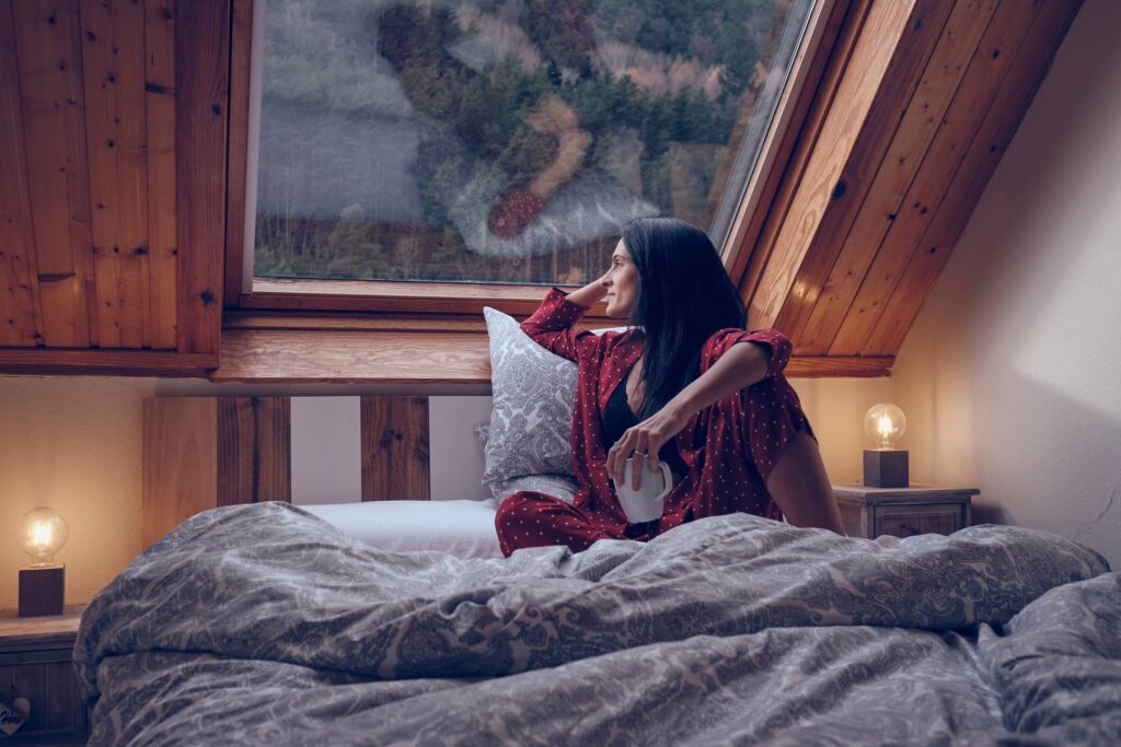 Donna in pigiamo a letto che guarda dalla finestra una foresta di pini e cerca di rilassarsi