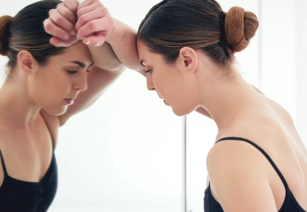 Donna con i capelli raccolti appoggiata a uno specchio, con la fronte poggiata sulle braccia e gli occhi chiusi, in un momento di stanchezza o tensione emotiva.