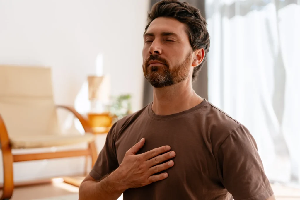 Uomo seduto in una stanza luminosa con gli occhi chiusi e una mano appoggiata sul petto mentre pratica un esercizio di respirazione.
