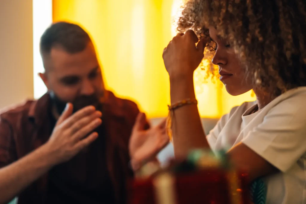 Donna con i ricci e sguardo preoccupato seduta davanti a un uomo che parla gesticolando; scena di conversazione intensa in un ambiente domestico con luce calda.